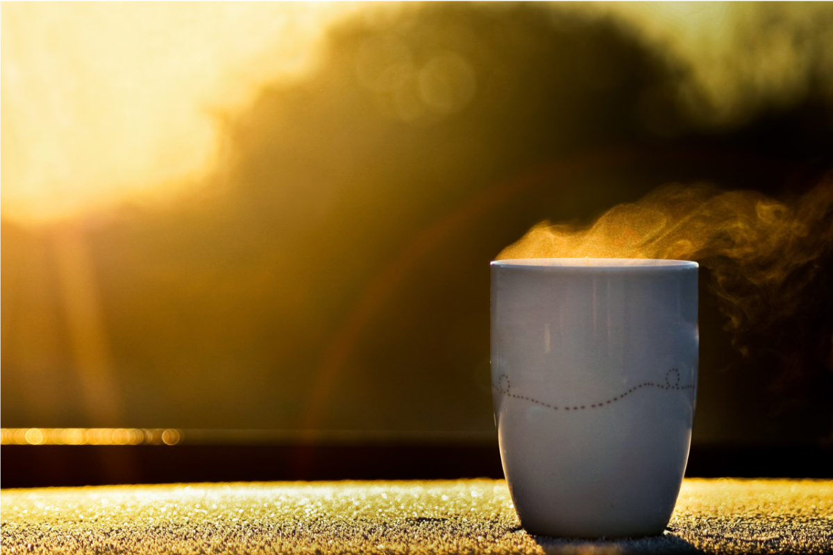 A cup of coffee on a table in front of a yellow morning sun, highlighting the steam