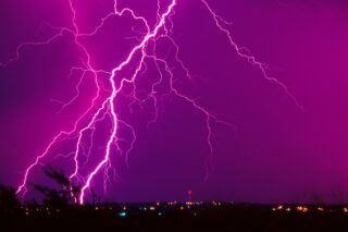 A multi-prongued lightning bolt against a purple night hitting a silhouetted city skyline dappled with lights.