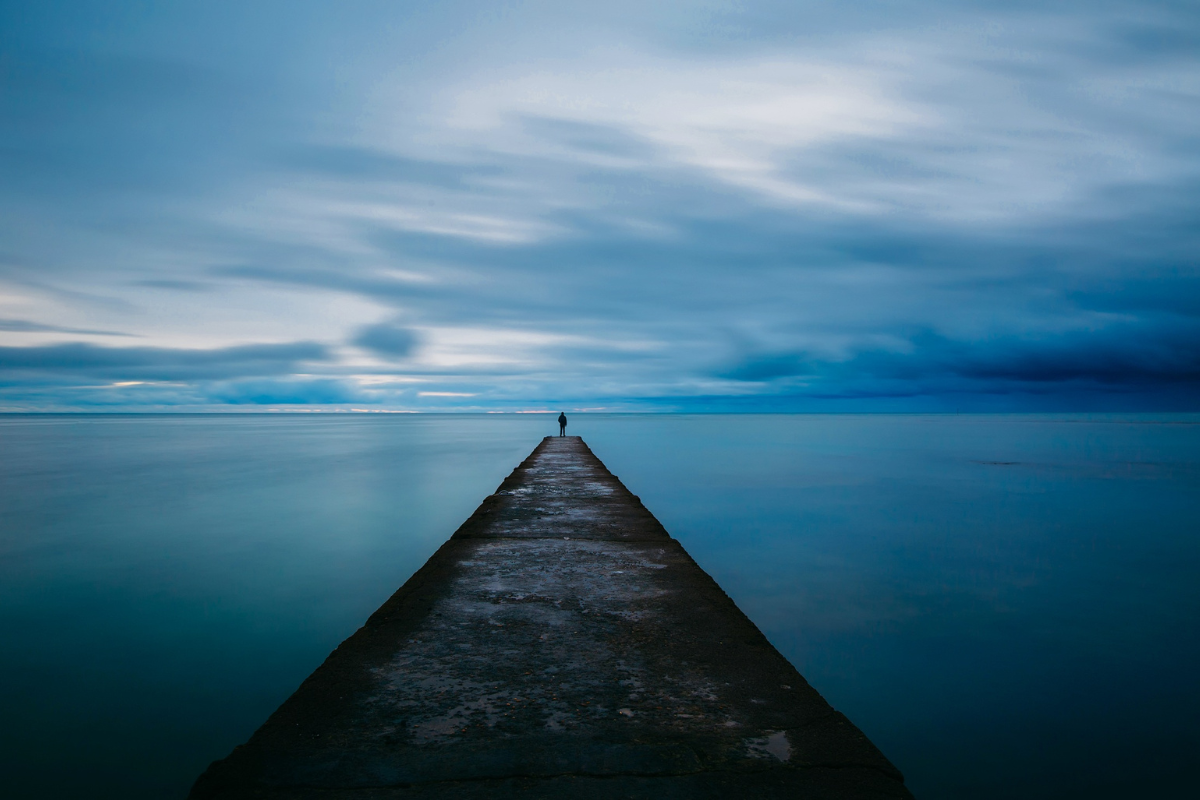 lonely – issue 73 – rough cut press A figure standing at the end of a long dock over water at dusk